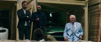 Movie still from “Collide” (2016), directed by Eran Creevy – A group of men standing next to each other in front of a car; Medium shot, Over the shoulder angle