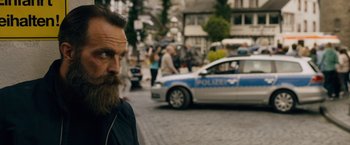 Movie still from “Collide” (2016), directed by Eran Creevy – A man with a long beard standing in front of a police car; Close Up shot, Over the shoulder angle