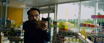 Movie still from “Collide” (2016), directed by Eran Creevy – A man holding a gun in front of a store window; Medium shot, Over the shoulder angle
