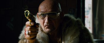 Movie still from “Collide” (2016), directed by Eran Creevy – An older man wearing a fur coat and glasses holding a gun; Close Up shot, Low angle