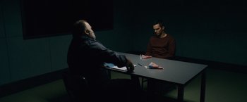 Movie still from “Collide” (2016), directed by Eran Creevy – Two men sitting at a table in a dark room; Medium shot, Over the shoulder angle