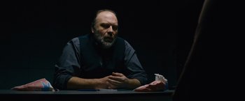 Movie still from “Collide” (2016), directed by Eran Creevy – A man sitting at a table in a dark room; Medium shot, Over the shoulder angle