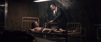 Movie still from “Colonia” (2015), directed by Florian Gallenberger – A man in a suit and tie standing over a woman in a bed; Medium shot, Low angle