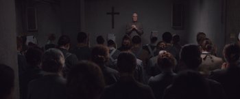Movie still from “Colonia” (2015), directed by Florian Gallenberger – A group of people sitting in front of a cross in a room; Wide shot, High angle