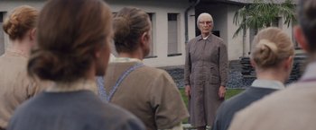 Movie still from “Colonia” (2015), directed by Florian Gallenberger – An old woman standing in front of a group of people; Medium shot, Over the shoulder angle