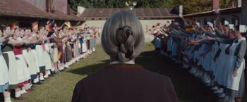 Movie still from “Colonia” (2015), directed by Florian Gallenberger – A woman with a long gray hair standing in front of a group of people; Medium shot, Over the shoulder angle