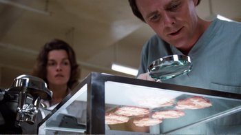 Movie still from “Coma” (1978), directed by Michael Crichton – A man and a woman looking through a magnifying glass at food; Close Up shot, Over the shoulder angle