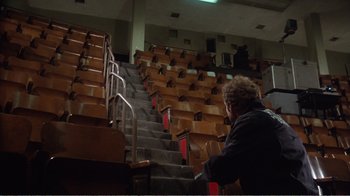 Movie still from “Coma” (1978), directed by Michael Crichton – A man sitting in front of a row of wooden chairs; Wide shot, High angle