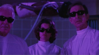 Movie still from “Coma” (1978), directed by Michael Crichton – A woman and two men wearing sunglasses in a purple room; Medium shot, Low angle