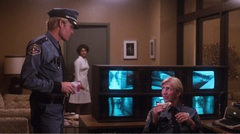 Movie still from “Coma” (1978), directed by Michael Crichton – A police officer is talking to a man in front of a television; Wide shot, Over the shoulder angle