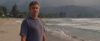 Movie still from “The Descendants” (2011), directed by Alexander Payne – A man standing on the beach with a surfboard in the background; Medium shot, Over the shoulder angle