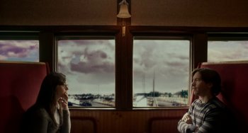Movie still from “Comet” (2014), directed by Sam Esmail – Two people sitting in front of two large windows on a train; Medium shot, Over the shoulder angle