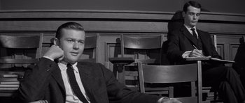 Movie still from “Compulsion” (1959), directed by Richard Fleischer – A black and white photo of a man in a suit and tie sitting in front of a bookshelf; Medium shot, Low angle
