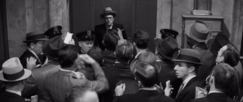 Movie still from “Compulsion” (1959), directed by Richard Fleischer – A group of men in suits and hats talking to each other; Medium shot, Over the shoulder angle