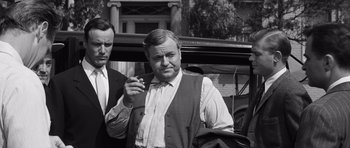 Movie still from “Compulsion” (1959), directed by Richard Fleischer – A man standing next to another man smoking a cigarette in front of an antique car; Medium shot, Over the shoulder angle