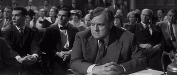 Movie still from “Compulsion” (1959), directed by Richard Fleischer – A man in a suit and tie sitting in front of a group of people; Medium shot, Over the shoulder angle