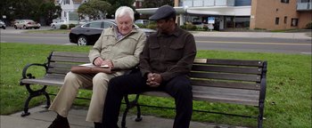 Movie still from “The Equalizer 2” (2018), directed by Antoine Fuqua – Two men sitting on a park bench , one of which is writing; Medium shot, Over the shoulder angle