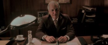 Movie still from “Confessions of a Dangerous Mind” (2002), directed by George Clooney – An older man sitting at a desk in a suit and tie; Medium shot, Low angle