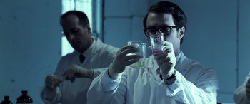 Movie still from “Confessions of a Dangerous Mind” (2002), directed by George Clooney – A man in a lab coat holding up a glass; Medium shot, Over the shoulder angle