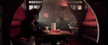 Movie still from “Confessions of a Dangerous Mind” (2002), directed by George Clooney – A woman sitting at a table holding a martini glass; Wide shot, High angle
