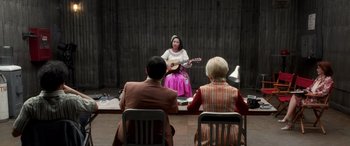 Movie still from “Confessions of a Dangerous Mind” (2002), directed by George Clooney – Three people sitting in chairs watching a woman play a guitar; Wide shot, Over the shoulder angle