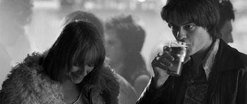 Movie still from “Control” (2007), directed by Anton Corbijn – A man and a woman are standing in front of a group of people; Medium shot, Over the shoulder angle