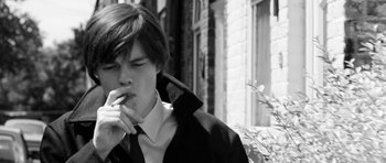 Movie still from “Control” (2007), directed by Anton Corbijn – A young man smoking a cigarette in front of a brick building; Close Up shot, Low angle