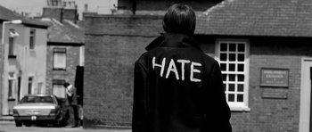 Movie still from “Control” (2007), directed by Anton Corbijn – A person wearing a jacket that says " hate " on the back of it; Medium shot, Over the shoulder angle