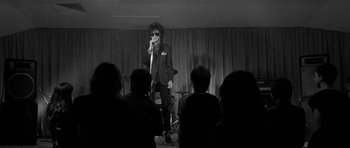 Movie still from “Control” (2007), directed by Anton Corbijn – A man in a black suit and sunglasses is on a microphone; Wide shot, Low angle