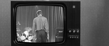 Movie still from “Control” (2007), directed by Anton Corbijn – A black and white photo of a man standing in front of a tv; Medium shot, Low angle