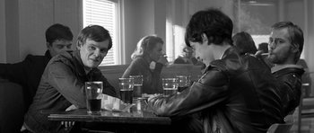 Movie still from “Control” (2007), directed by Anton Corbijn – A group of people sitting at a table with drinks; Medium shot, Over the shoulder angle