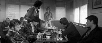 Movie still from “Control” (2007), directed by Anton Corbijn – A group of people sitting at a table with drinks; Medium shot, Over the shoulder angle