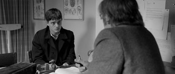 Movie still from “Control” (2007), directed by Anton Corbijn – A young man sitting in front of another man in front of a mirror; Medium shot, Over the shoulder angle