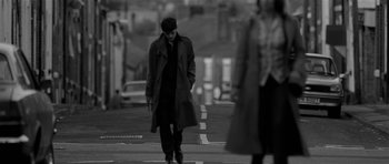 Movie still from “Control” (2007), directed by Anton Corbijn – A man in a trench coat and hat is crossing the street; Wide shot, Over the shoulder angle