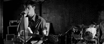 Movie still from “Control” (2007), directed by Anton Corbijn – A man holding a guitar in front of a drum set; Medium shot, Low angle