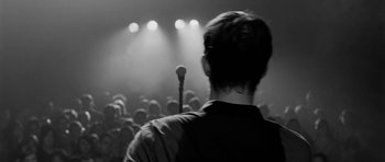 Movie still from “Control” (2007), directed by Anton Corbijn – A man standing in front of a microphone on a stage; Medium shot, Over the shoulder angle
