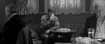Movie still from “Control” (2007), directed by Anton Corbijn – A man sitting at a table drinking from a glass; Medium shot, High angle
