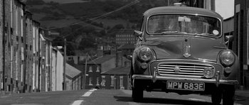 Movie still from “Control” (2007), directed by Anton Corbijn – An old car is parked on the side of the road; Wide shot, Low angle