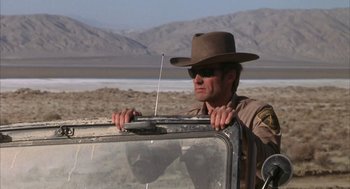 Movie still from “Coogan's Bluff” (1968), directed by Don Siegel – A man in a hat and sunglasses standing in the back of a truck; Medium shot, Low angle