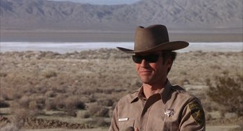 Movie still from “Coogan's Bluff” (1968), directed by Don Siegel – A man wearing a hat and sunglasses standing in front of a field; Medium shot, Over the shoulder angle