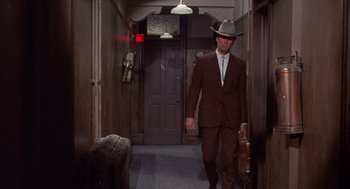 Movie still from “Coogan's Bluff” (1968), directed by Don Siegel – A man in a suit and hat walking down a hall way; Wide shot, Over the shoulder angle