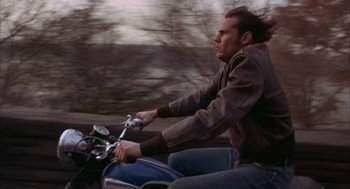Movie still from “Coogan's Bluff” (1968), directed by Don Siegel – A man riding on the back of a blue motorcycle; Medium shot, Low angle