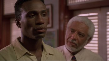 Movie still from “Cool Runnings” (1993), directed by Jon Turteltaub – A man standing next to another man in a room; Close Up shot, Over the shoulder angle