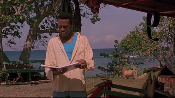 Movie still from “Cool Runnings” (1993), directed by Jon Turteltaub – A man standing in front of the ocean holding a book; Medium shot, Low angle