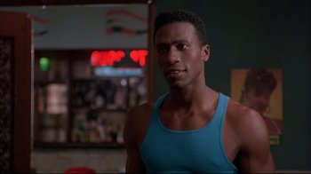 Movie still from “Cool Runnings” (1993), directed by Jon Turteltaub – A man in a blue tank top is standing in front of a bar; Close Up shot, Over the shoulder angle