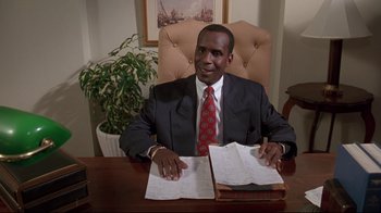 Movie still from “Cool Runnings” (1993), directed by Jon Turteltaub – A man sitting at a desk in front of papers; Medium shot, Low angle