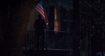 Movie still from “The Front Runner” (2018), directed by Jason Reitman – A man standing on a porch holding an american flag at night; Wide shot, Low angle