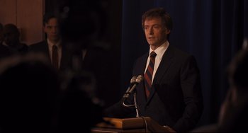 Movie still from “The Front Runner” (2018), directed by Jason Reitman – A man wearing a suit and tie standing in front of a microphone; Medium shot, Over the shoulder angle