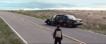Movie still from “Cop Car” (2015), directed by Jon Watts – A man standing on the side of a road near a police car; Wide shot, High angle