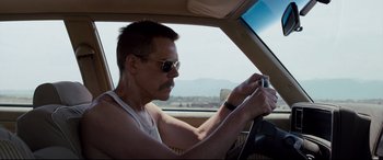 Movie still from “Cop Car” (2015), directed by Jon Watts – A man driving a car while holding a steering wheel; Close Up shot, Over the shoulder angle